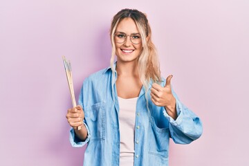 Beautiful young blonde woman holding paintbrushes smiling happy and positive, thumb up doing excellent and approval sign