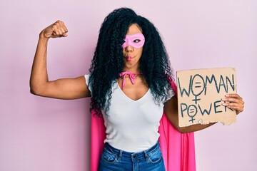 Middle age african american woman wearing super hero costume holding woman power banner making fish face with mouth and squinting eyes, crazy and comical.