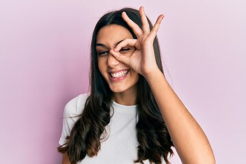 Young hispanic woman wearing casual white t shirt smiling happy doing ok sign with hand on eye looking through fingers