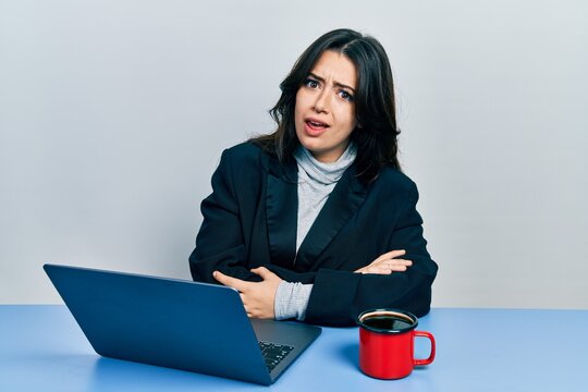 Beautiful hispanic business woman with arms crossed gesture at the office in shock face, looking skeptical and sarcastic, surprised with open mouth