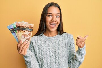 Beautiful hispanic woman holding canadian dollars pointing thumb up to the side smiling happy with...