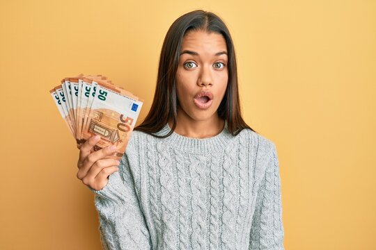 Beautiful hispanic woman holding bunch of 50 euro banknotes scared and amazed with open mouth for surprise, disbelief face