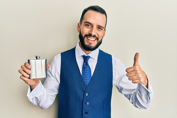 Young man with beard drinking whiskey from flask smiling happy and positive, thumb up doing excellent and approval sign