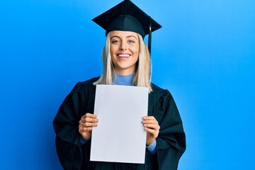 Beautiful blonde woman wearing graduation cap and ceremony robe holding blank banner smiling and...