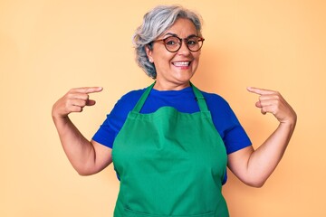 Senior hispanic woman wearing apron and glasses looking confident with smile on face, pointing...