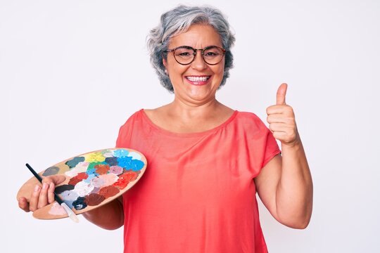 Senior hispanic grey- haired woman holding paintbrush and palette smiling happy and positive, thumb up doing excellent and approval sign