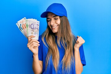 Young hispanic woman wearing delivery uniform and cap holding singapore dollars screaming proud, celebrating victory and success very excited with raised arm