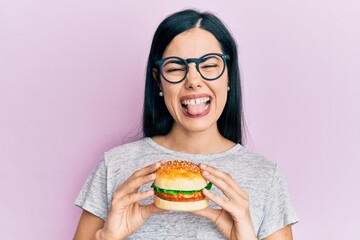 Beautiful young woman eating tasty hamburger sticking tongue out happy with funny expression.