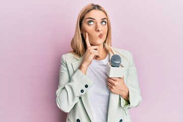 Young caucasian woman holding reporter microphone serious face thinking about question with hand on...