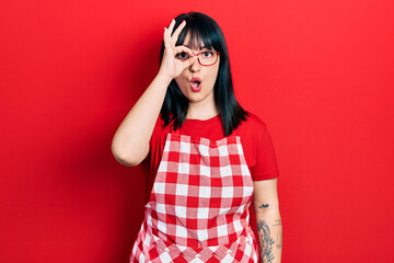 Young hispanic woman wearing cook apron and glasses doing ok gesture shocked with surprised face, eye looking through fingers. unbelieving expression.