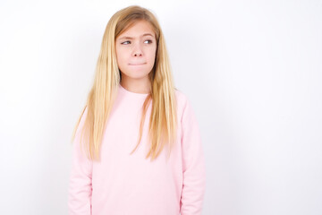Amazed puzzled beautiful caucasian little girl wearing pink hoodie over white background, curves lips and has worried look, sees something awful in front.