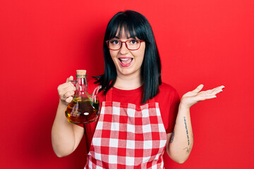 Young hispanic woman wearing cook apron holding olive oil can celebrating achievement with happy...