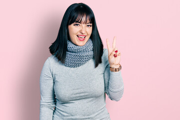 Young hispanic plus size woman wearing winter scarf smiling with happy face winking at the camera doing victory sign with fingers. number two.