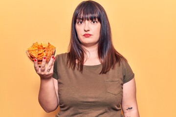 Young plus size woman holding nachos potato chips thinking attitude and sober expression looking self confident