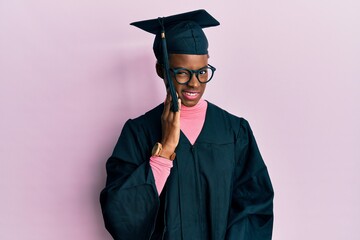Young african american girl wearing graduation cap and ceremony robe touching mouth with hand with...