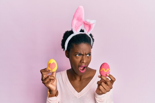 Young african american girl wearing cute easter bunny ears holding painted eggs angry and mad screaming frustrated and furious, shouting with anger. rage and aggressive concept.