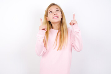 Successful friendly looking beautiful caucasian little girl wearing pink hoodie over white background exclaiming excitedly, pointing both index fingers up, indicating something.