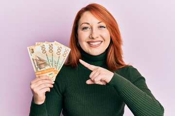 Beautiful redhead woman holding 500 norwegian krone banknotes smiling happy pointing with hand and...