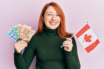 Beautiful redhead woman holding canadian dollars and canada flag winking looking at the camera with...