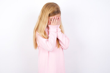 Sad beautiful caucasian little girl wearing pink hoodie over white background covering face with hands and crying.