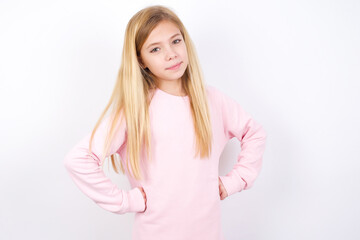 Funny frustrated beautiful caucasian little girl wearing pink hoodie over white background holding hands on waist and silly looking at awkward situation.