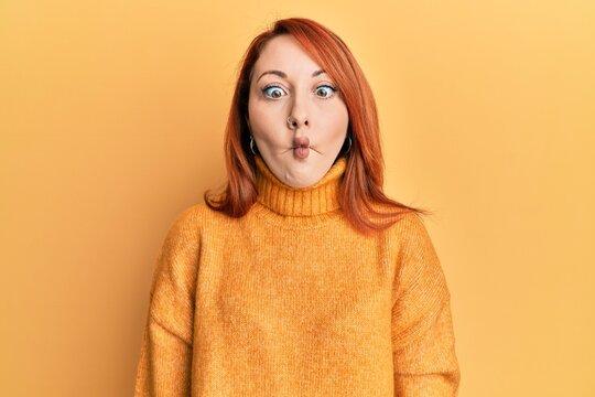 Beautiful redhead woman wearing casual winter sweater over yellow background making fish face with lips, crazy and comical gesture. funny expression.