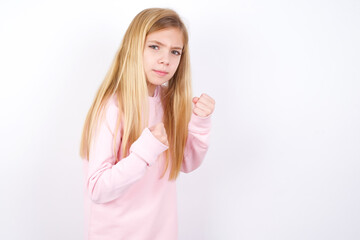 Portrait of attractive beautiful caucasian little girl wearing pink hoodie over white background holding hands in front of him in boxing position going to fight.