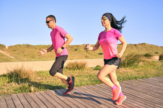 Active couple running during a warm summer day. Wearing pink shirts and short pants. Both are wearing sunglasses. Wooden pathway surrounded by grass.