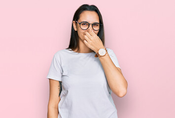 Young hispanic woman wearing casual white t shirt smelling something stinky and disgusting,...