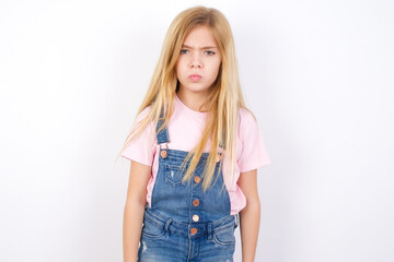 Obraz premium Offended dissatisfied beautiful caucasian little girl wearing jeans overall over white background with moody displeased expression at camera being disappointed by something