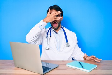 Handsome hispanic man wearing doctor uniform working at the clinic peeking in shock covering face and eyes with hand, looking through fingers afraid