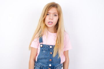 Obraz premium Portrait of dissatisfied beautiful caucasian little girl wearing jeans overall over white wall smirks face, purses lips and looks with annoyance at camera, discontent hearing something unpleasant