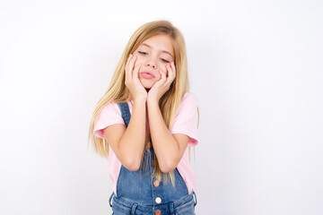 Fototapeta premium beautiful caucasian little girl wearing jeans overall over white background keeps hands on cheeks has bored displeased expression. Stressed hopeless model
