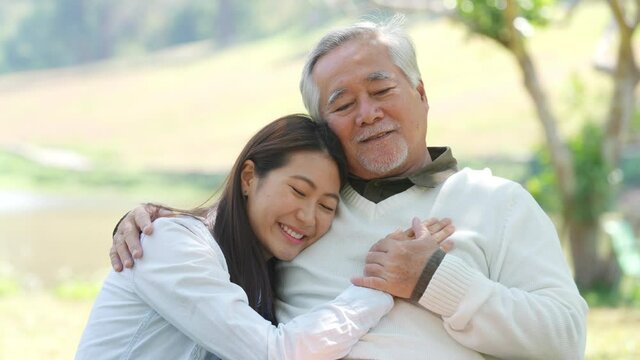 4K Asian woman caring and hugging senior man grandfather sitting on outdoor chair in the park. Elderly retired male relax and enjoy outdoor activity together with daughter. Family relationship concept