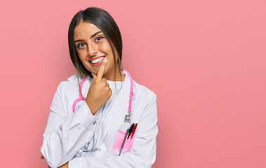 Beautiful hispanic woman wearing doctor uniform and stethoscope looking confident at the camera smiling with crossed arms and hand raised on chin. thinking positive.