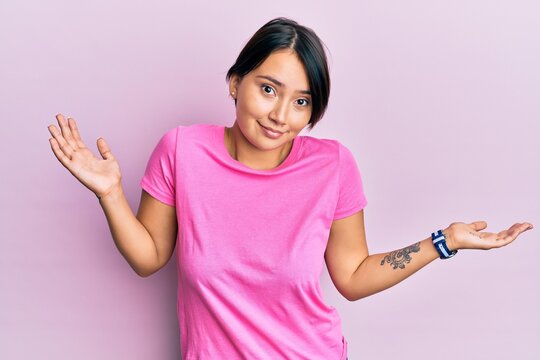 Beautiful young woman with short hair wearing pink tshirt over pink background clueless and confused with open arms, no idea and doubtful face.