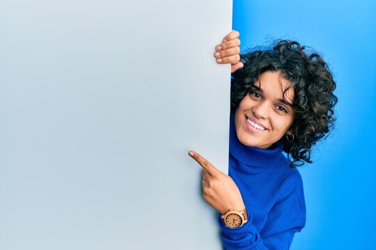 Young hispanic woman with curly hair holding blank empty banner smiling happy pointing with hand and finger