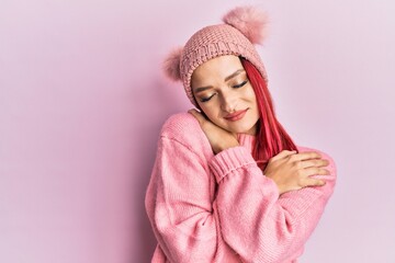 Young caucasian woman wearing wool sweater and winter hat hugging oneself happy and positive, smiling confident. self love and self care