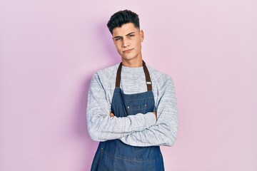 Young hispanic man wearing apron skeptic and nervous, disapproving expression on face with crossed...