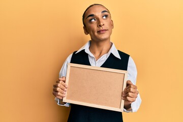 Hispanic man wearing make up and long hair holding empty corkboard smiling looking to the side and staring away thinking.