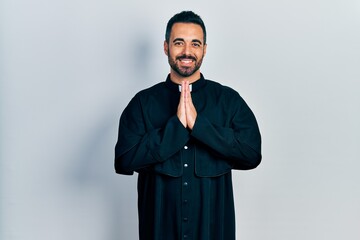 Handsome hispanic man with beard wearing catholic priest robe praying with hands together asking...