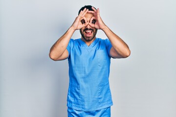 Fototapeta premium Handsome hispanic man with beard wearing blue male nurse uniform doing ok gesture like binoculars sticking tongue out, eyes looking through fingers. crazy expression.