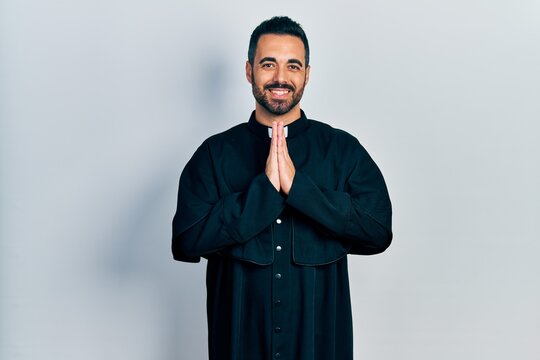 Handsome hispanic man with beard wearing catholic priest robe praying with hands together asking for forgiveness smiling confident.