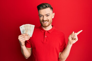 Young redhead man holding 100 danish krone banknotes smiling happy pointing with hand and finger to...