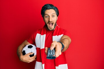 Young hispanic man football hooligan holding ball using tv control celebrating crazy and amazed for...