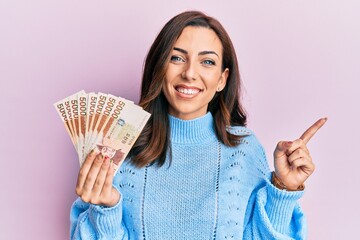 Young brunette woman holding 5000 south korean won banknotes smiling happy pointing with hand and finger to the side