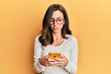 Young brunette woman using smartphone over yellow background relaxed with serious expression on...