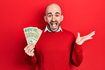 Young bald man holding argentine pesos banknotes celebrating achievement with happy smile and...