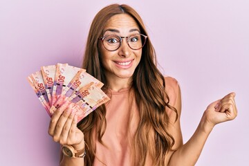 Young brunette woman holding south african rand banknotes screaming proud, celebrating victory and success very excited with raised arm