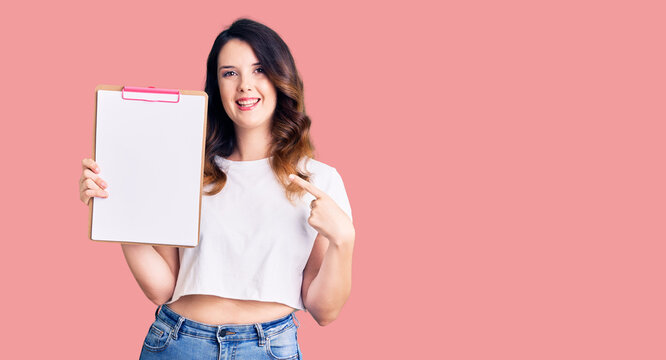 Beautiful young brunette woman holding clipboard with blank space smiling happy pointing with hand and finger